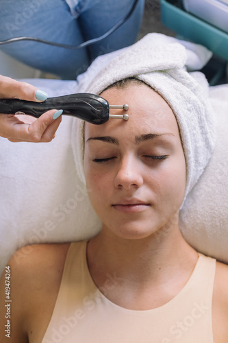 Top view of crop unrecognizable female cosmetologist doing rejuvenate facial procedure with microcurrent device for graceful lady lying on couch with closed eyes photo