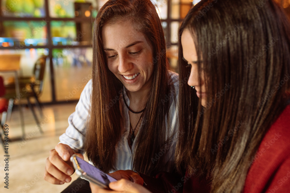 Young cheerful lesbian couple sitting at table in cafe and using smartphone while spending weekend together Young cheerful lesbian couple sitting at table in cafe and using smartphone while spending weekend together