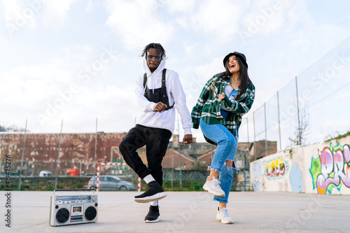 Young glad stylish multiracial partners dancing hip hop on pavement near retro tape recorder under cloudy sky in town