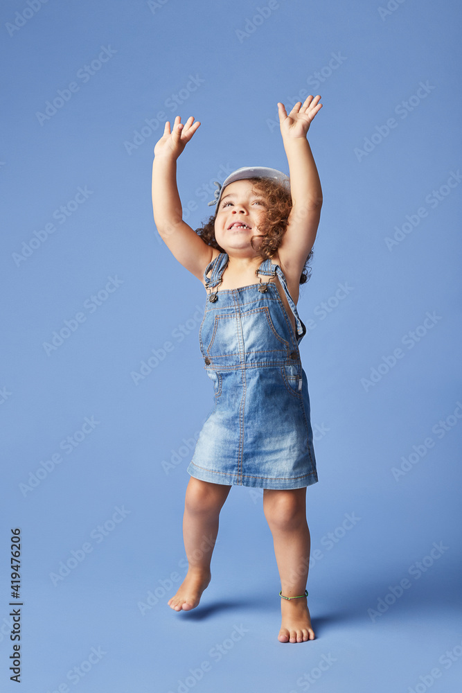 Charming barefoot child in denim dress and hat with curly hair looking up with arms raised while dancing on blue background Charming barefoot child in denim dress and hat with curly hair looking up with arms raised while dancing on blue background