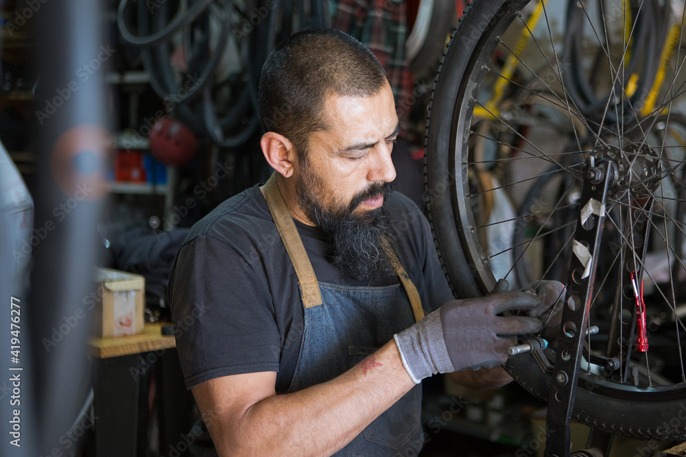 Serious adult man in apron and gloves repairing wheel of bike in modern garage Serious adult man in apron and gloves repairing wheel of bike in modern garage