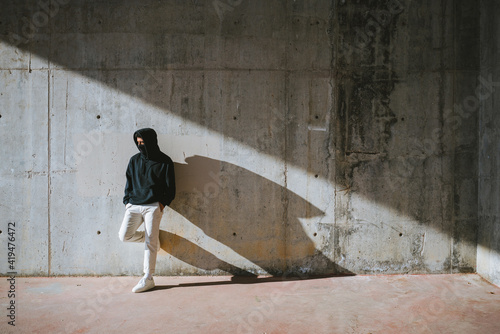 Unrecognizable young male in hoodie and mask standing on street near concrete wall
