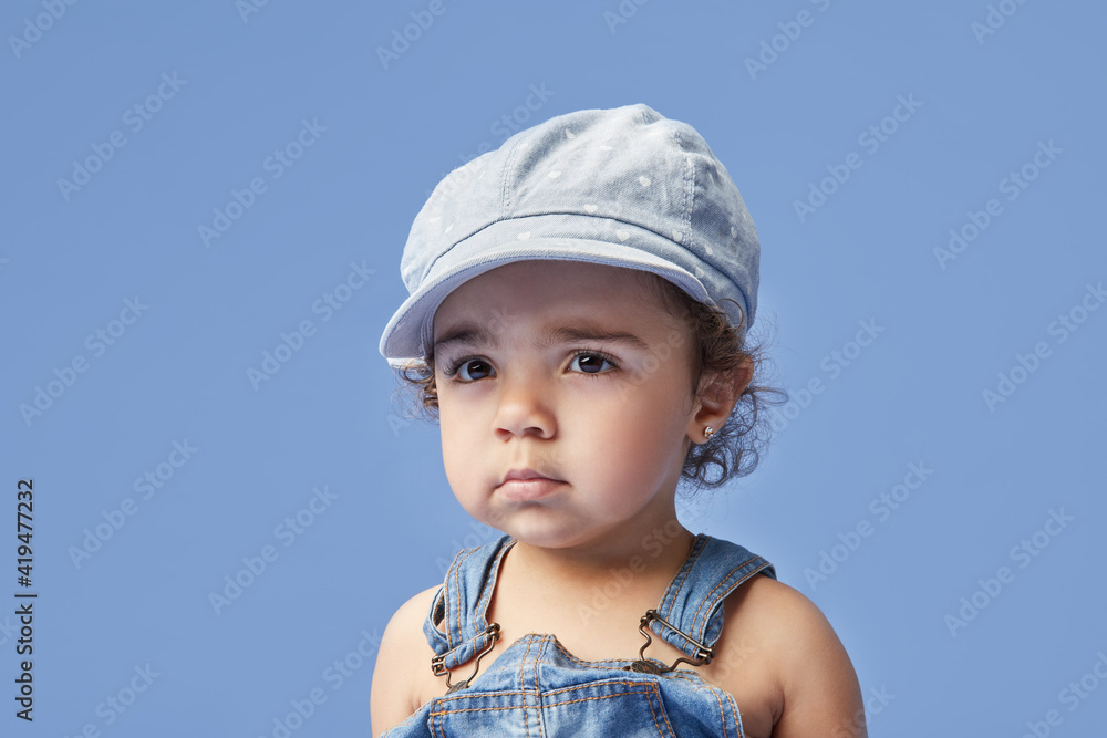 Adorable upset little kid with brown eyes wearing casual clothes and hat against blue background looking away Adorable upset little kid with brown eyes wearing casual clothes and hat against blue background looking away