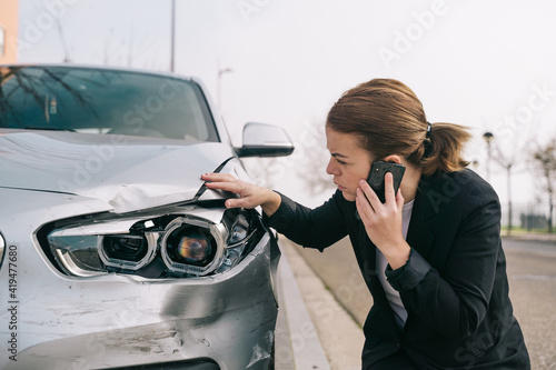 Upset young female driver in casual outfit having phone conversation on road and inspecting damages on modern car after crash
