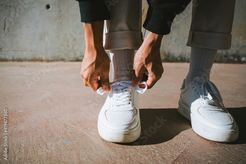 From above of crop unrecognizable trendy guy tying laces of stylish sneakers while standing on street on sunny day