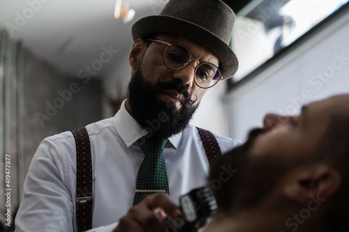 Stylish mature dandy ethnic male beauty master cutting mustache of bearded client using trimmer in barbershop