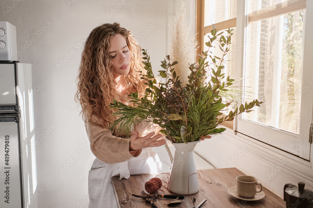 Gentle female standing at table with bunch of fresh green plants in vase at home Gentle female standing at table with bunch of fresh green plants in vase at home