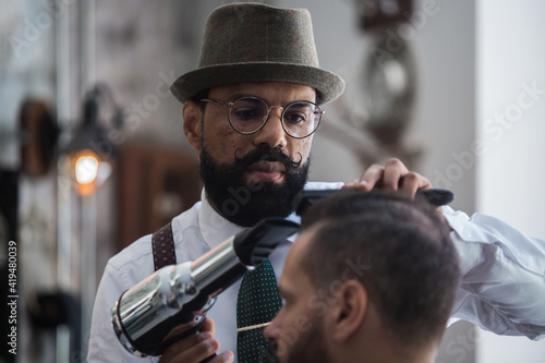 Professional mature ethnic male beauty master drying hair of crop male client using hairdryer in barbershop photo