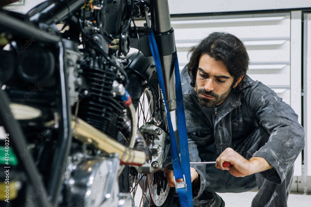 Busy male technician in uniform repairing wheel of shiny motorbike with screwdriver in workshop