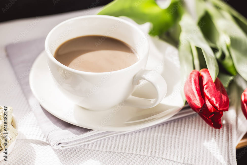 Obraz premium Close-up morning cup of coffee with milk, candy and tulip on the table. Light rays fall on the table