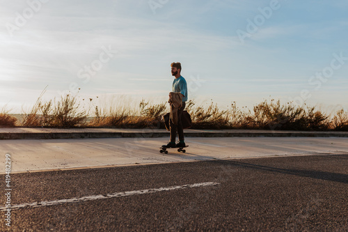 Full body young bearded male riding skateboard with bag and jacket in hand along pavement near asphalt road photo