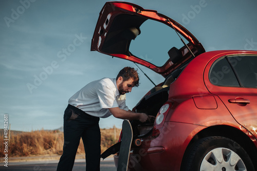 Side view of young bearded male driver with opened car trunk getting dressed