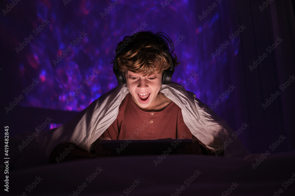 Excited teenage boy in headphones sitting on bed in dark room with glowing lights while smiling and watching video on tablet