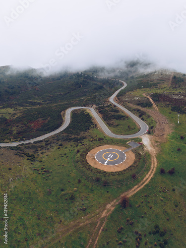 Spectacular drone view of curved serpent road near round concrete helipad on grassy rural valley on summer day running through green hills in highlands