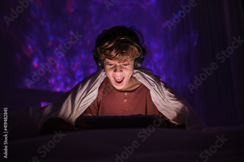 Excited teenage boy in headphones sitting on bed in dark room with glowing lights while smiling and watching video on tablet