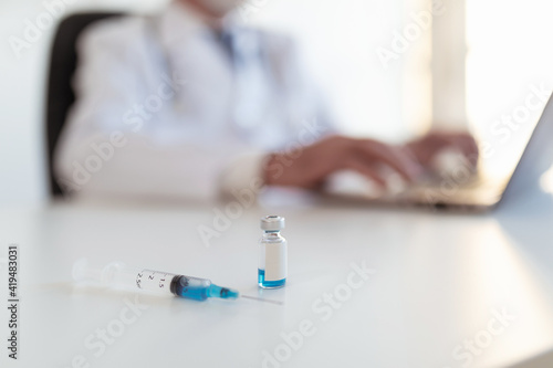 Crop anonymous doctor working on netbook on table with injector and small bottle with blue substance in clinic