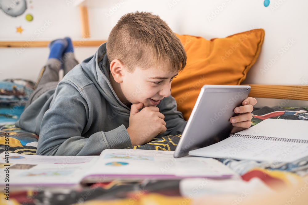 Side view of smiling teenage boy lying on bed and talking to classmates via tablet during online lesson at home Side view of smiling teenage boy lying on bed and talking to classmates via tablet during online lesson at home