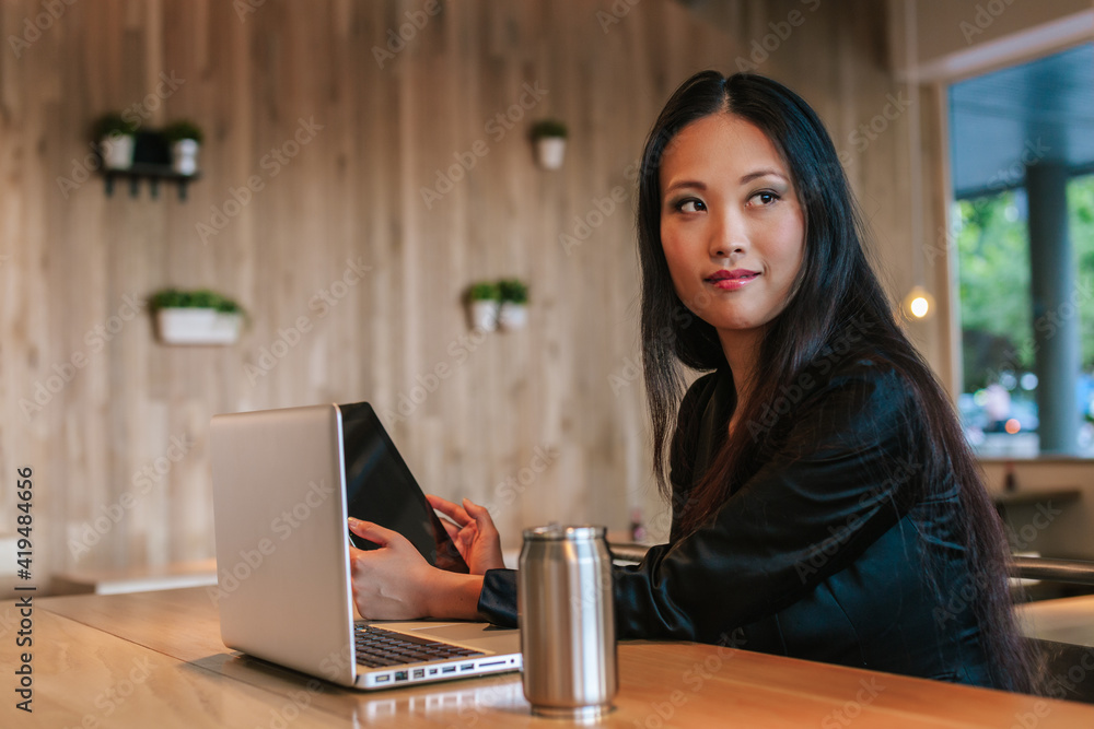 Content ethnic female entrepreneur sitting at table with tablet and laptop while working on project and looking away Content ethnic female entrepreneur sitting at table with tablet and laptop while working on project and looking away