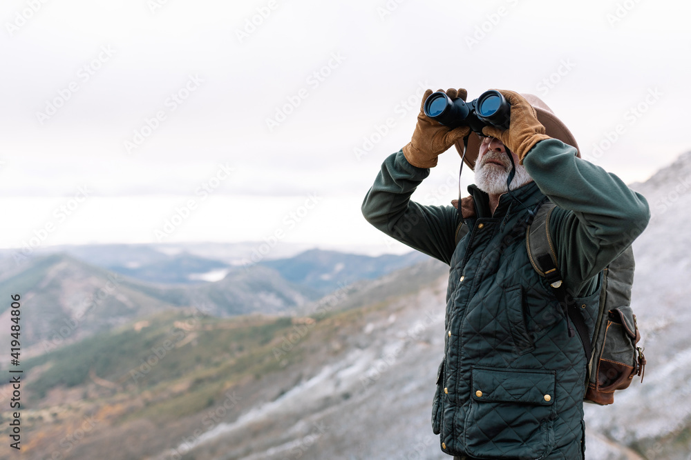 Senior male traveler in outerwear looking through binoculars while standing in highland terrain and admiring picturesque landscape during winter holidays in Caceres Senior male traveler in outerwear looking through binoculars while standing in highland terrain and admiring picturesque landscape during winter holidays in Caceres