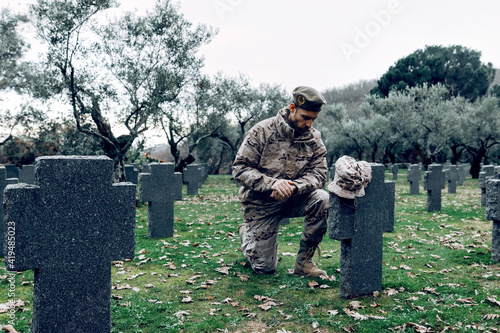 Full body sorrowful soldier in camouflage outfit kneeling down in front of grave in military cemetery on early autumn day