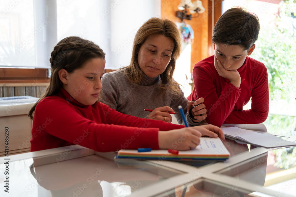 Teenage schoolgirl sitting at table with mother and brother and writing in copybook while doing homework together at home Teenage schoolgirl sitting at table with mother and brother and writing in copybook while doing homework together at home