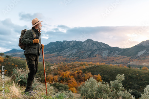 Side view of elderly male wanderer with wooden stick standing on hill and admiring scenic view of highlands in Caceres