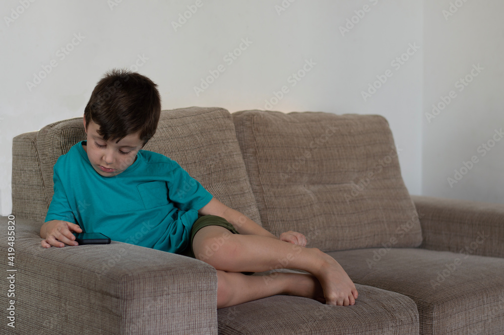 Cute little boy barefoot using smartphone, looking at screen, sitting on cozy couch in living room Cute little boy barefoot using smartphone, looking at screen, sitting on cozy couch in living room