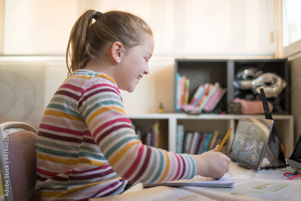 Schoolgirl talking on video chat on tablet while sitting at table at home and having online lesson Schoolgirl talking on video chat on tablet while sitting at table at home and having online lesson