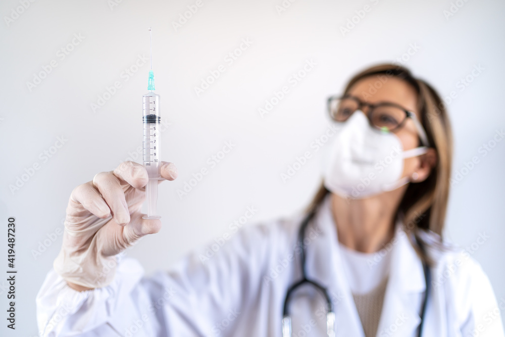 Female medic in protective mask and with vaccine from coronavirus in syringe standing in bright medical room of hospital Female medic in protective mask and with vaccine from coronavirus in syringe standing in bright medical room of hospital