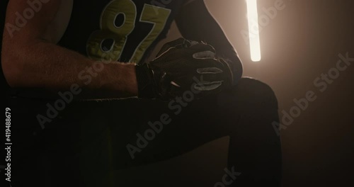 Crop rugby player cracking knuckles in dark studio. Crop unrecognizable male American football player in uniform and gloves sitting in dark studio and cracking knuckles