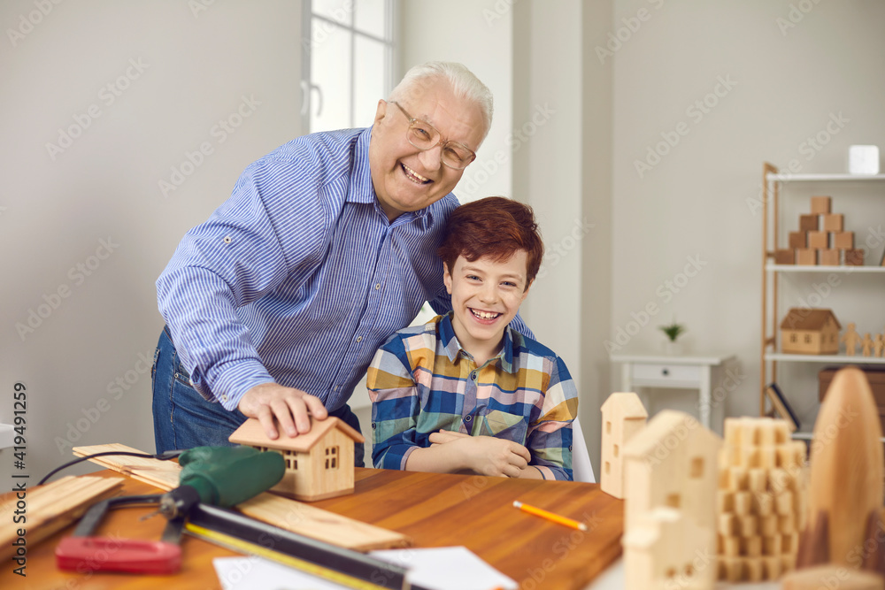 Obraz premium Experienced old carpenter teaches grandchild new handwork skills. Child and grandfather making wooden toy houses together. Happy smiling senior man with teen grandson having fun in carpentry workshop