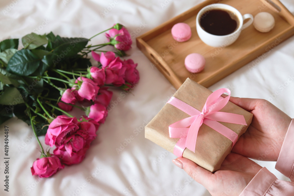 Woman's hand holding a gift. Cozy holiday morning composition with roses and cup of coffee with macaroons on wooden tray Woman's hand holding a gift. Cozy holiday morning composition with roses and cup of coffee with macaroons on wooden tray
