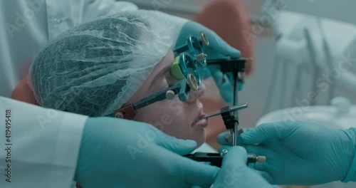 Close-up a female patient in disposable hat is sitting in the dentist chair, she is wearing one part of axiograph in front of her face and two dentists in disposable blue gloves. photo