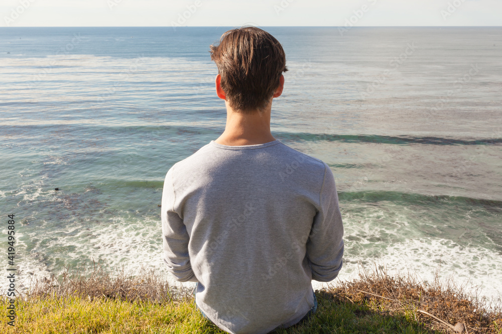 Stress-free wellness. Relaxed young man alone sitting on top a cliff facing the ocean view. Stress-free wellness. Relaxed young man alone sitting on top a cliff facing the ocean view.