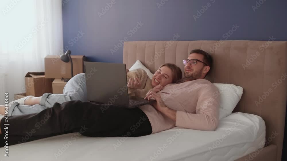 Young couple relaxes after moving in a new home. Couple lays on bed and looks at notebook screen Young couple relaxes after moving in a new home. Couple lays on bed and looks at notebook screen