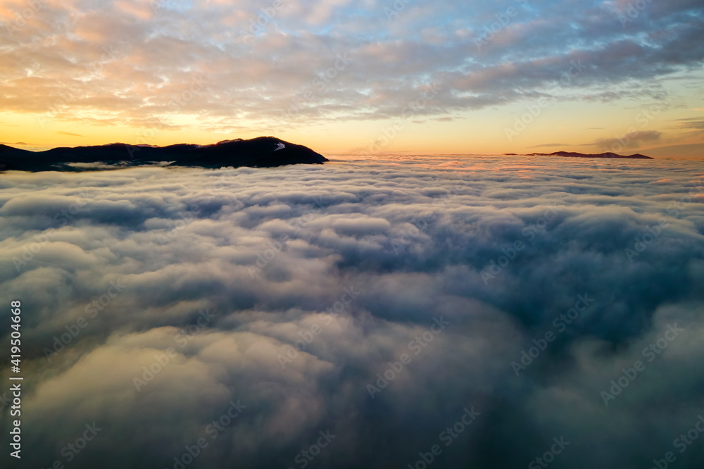 Aerial view of vibrant sunrise over white dense clouds with distant dark mountains on horizon.