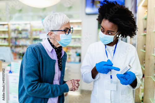 Professional Black female pharmacist talking with senior customer in modern drugstore. They are wearing face protective masks for protection from Coronavirus. photo