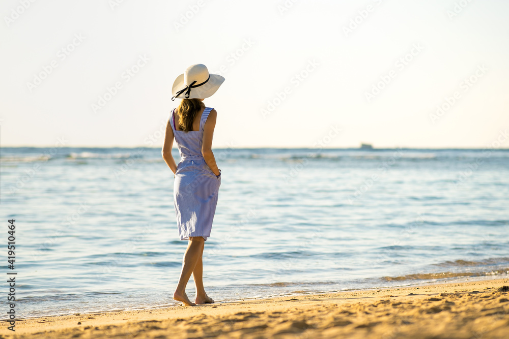 Young woman in straw hat and a dress walking alone on empty sand beach at sea shore. Lonely tourist girl looking at horizon over calm ocean surface on vacation trip.