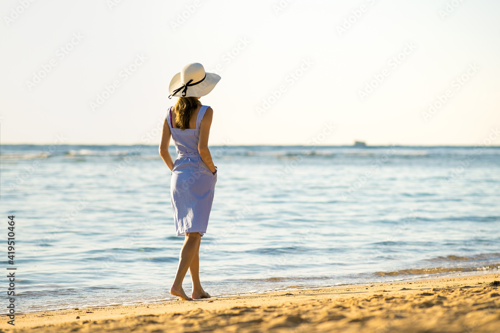 Young woman in straw hat and a dress walking alone on empty sand beach at sea shore. Lonely tourist girl looking at horizon over calm ocean surface on vacation trip. Young woman in straw hat and a dress walking alone on empty sand beach at sea shore. Lonely tourist girl looking at horizon over calm ocean surface on vacation trip.