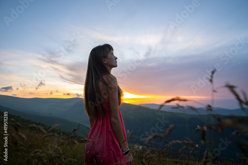 Young woman in red dress walking on grass field on a windy evening in autumn mountains.