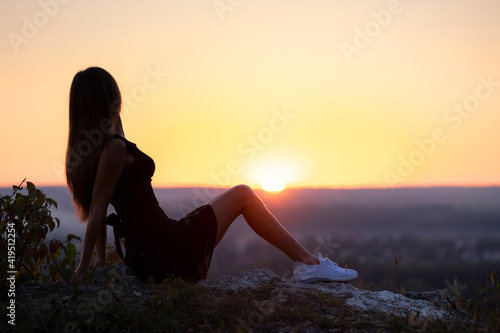 Young elegant woman in black short dress and white sneaker shoes sitting on a rock relaxing outdoors at summer evening. Fashionable lady enjoying warm sunset in nature.