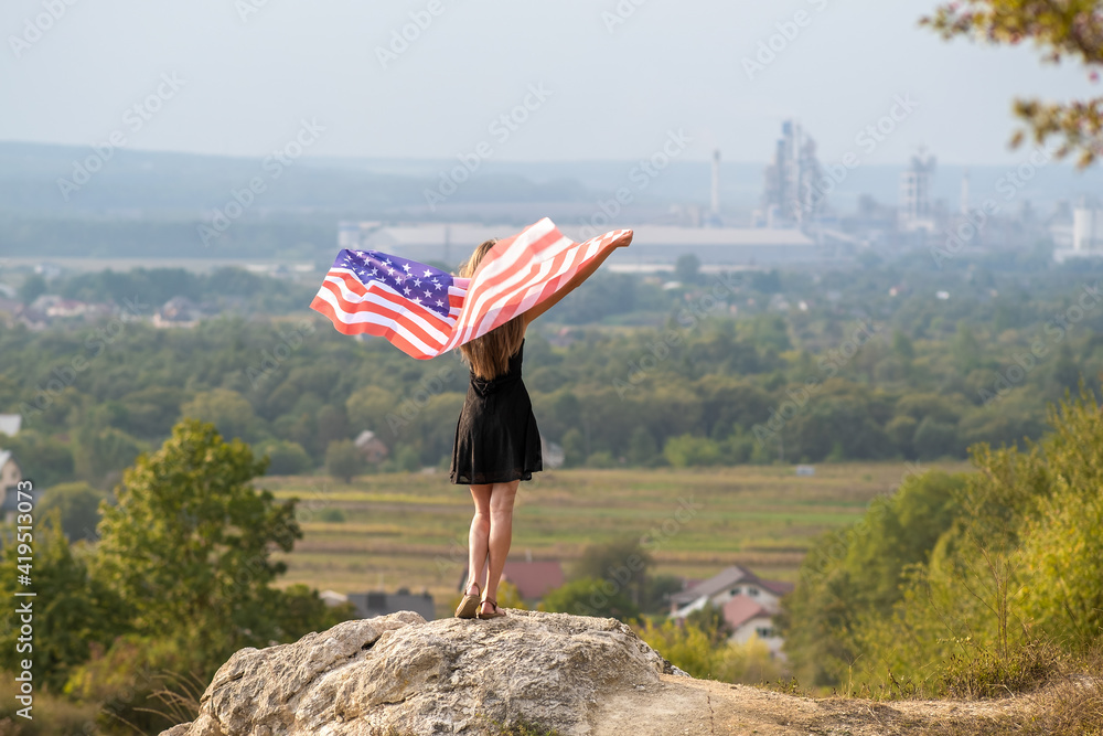 Obraz premium Young happy american woman with long hair raising up waving on wind USA national flag in her hands relaxing outdoors enjoying warm summer day.