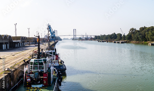 Two tugboats moored in the Guadalquivir river next to the warehouses and industrial units of the port of Seville on a sunny day (Andalusia, Spain). Views of the Centennial Bridge in the background.