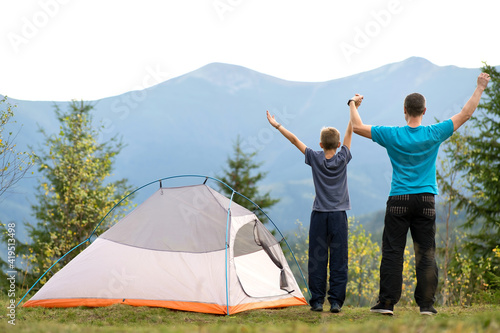 Young dad and his child son standing near camping tent with raised hands while hiking together in summer mountains. Active family travelling concept.