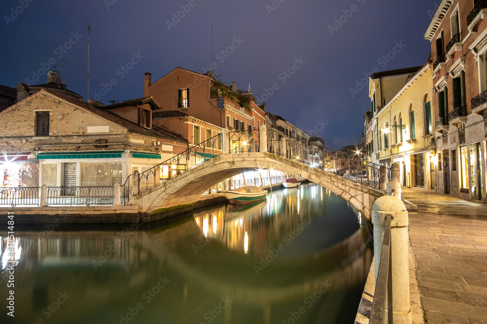Evening view of illuminated old architecture, floating boats and light reflections in canals water in Venice, Italy.