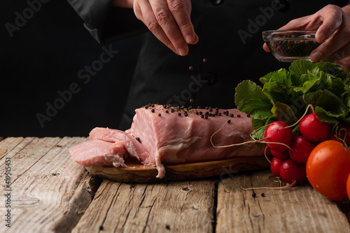 The chef sprinkles pork meat with pepper for cooking on the background of vegetables. On a black background.Butcher shop.Recipes with meat