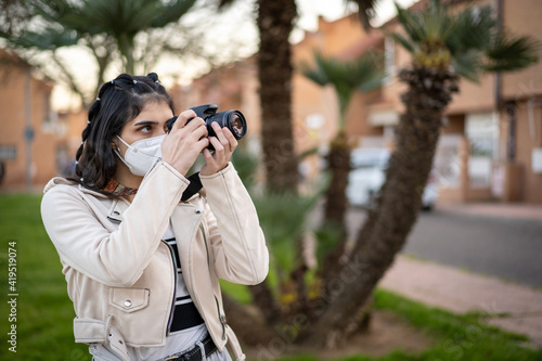 Joven mujer fotógrafa con una cámara réflex negra haciendo una foto al atardecer en un paisaje urbano, con cazadora blanca, camiseta de rayas, en un parque © Alex Visualnue
