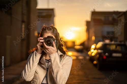 Joven mujer fotógrafa con una cámara réflex negra haciendo una foto al atardecer en un paisaje urbano, con cazadora blanca, camiseta de rayas, al atardecer photo