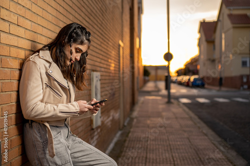 Mujer joven de pelo negro caucásica con camiseta de rayas negras, cazadora blanca, y pantalon vaquero, usando su smartphone,e scribiendo, apoyada en una pader de ladrillo en la calle al atardecer photo