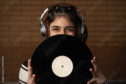 Mujer joven de pelo negro caucásica con auriculares y un disco de vinilo negro tapando su cara, enseñando sus ojos, sonriendo